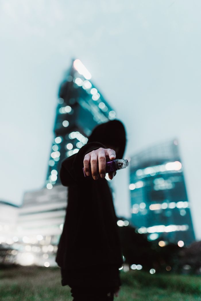 Mysterious hooded figure holding vape with skyscrapers in the background, evocative city vibe.