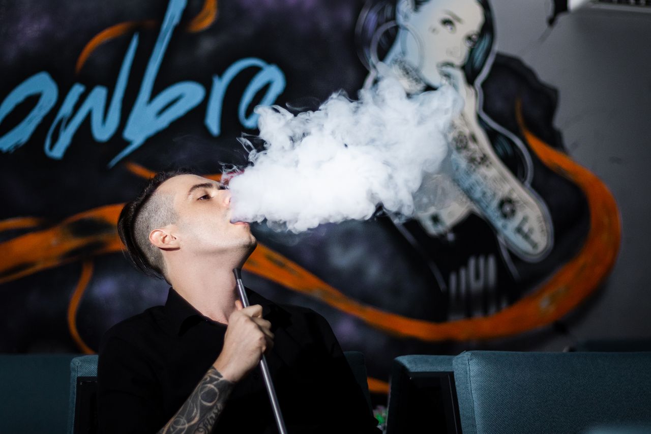 A man with tattoos vapes indoors, creating a smoke cloud against a mural backdrop.