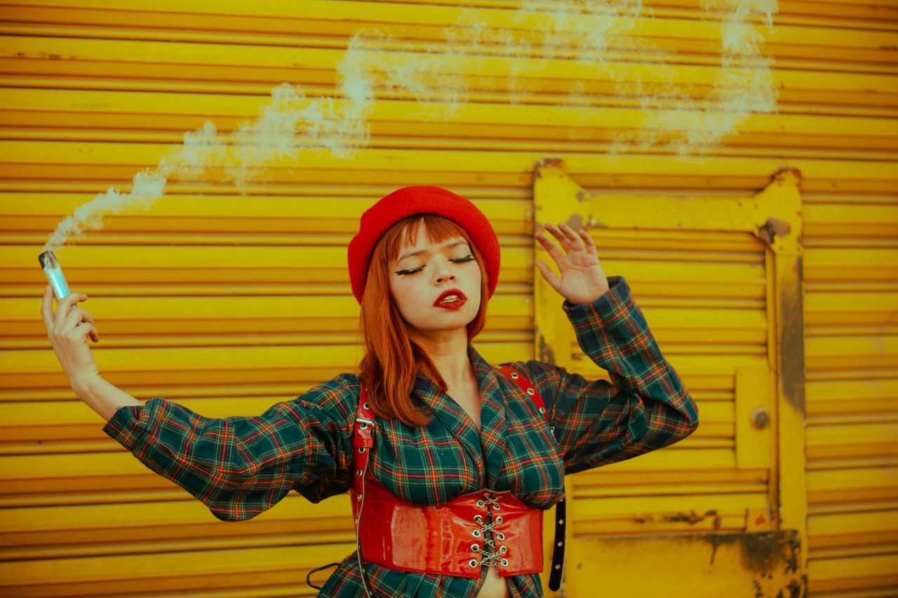 Stylish woman with red hat and vape against a vibrant yellow background.